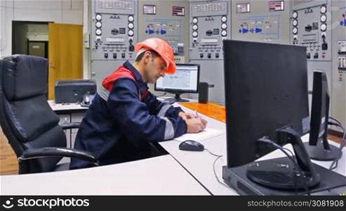 engineer sits at table and writes in log near main control panel of gas compressor station, closeup