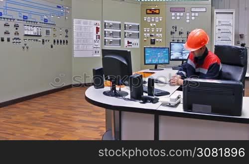 engineer sits at table and writes registry near main control panel of gas compressor station