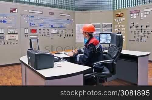 engineer sits at table in front of main control panel for gas compressor station and writes registry