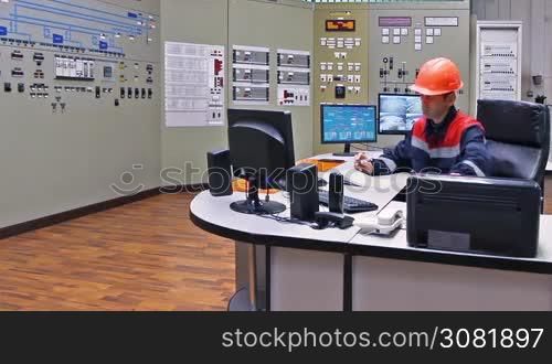 engineer takes phone and speaks while sitting at table on main control panel of gas compressor station