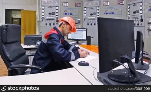 engineer writes down log and looks into monitor near main control panel of gas compressor station, closeup