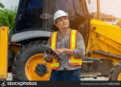 Engineering wearing a white safety helmet standing in front of the backhoe Looking at home construction work And use the tablet to check the blueprint with construction