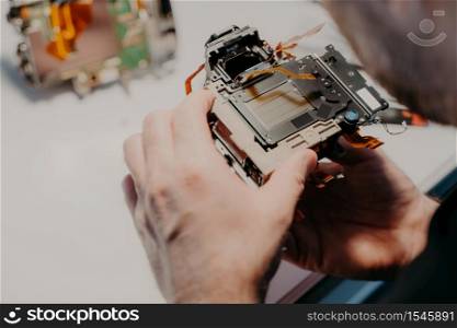 Engineers hands holds broken photo camera, poses against workspace, repairs digital device or professional dslr camera. Professional specialist with disassembled equipment