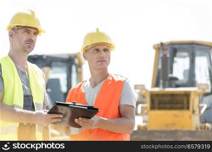 Engineers looking away while holding clipboard at construction site against clear sky