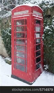 English red telephone box in winter snow against an ivy clad Cotswold stone wall.