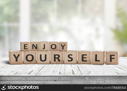 Enjoy yourself wriiten on wooden blocks in a bright room. Cubes with a positive phrase on a white desk in a living room with green plants.