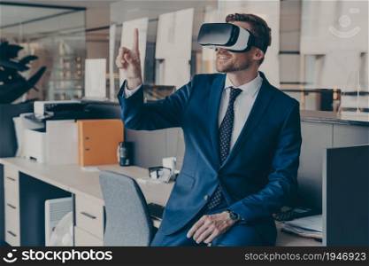 Enjoyed young businessman pointing with forefinger up in air with virtual reality glasses on head, while sitting on table in office with VR headset. Innovative gadget for business 3d visualizations. Enjoyed young businessman pointing with forefinger up in air with virtual reality glasses on head