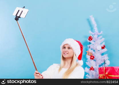 Enjoying christmas gifts. Woman in santa hat taking picture of herself using selfie stick. Indoor shot on blue background. Girl in santa hat taking picture of herself using selfie stick