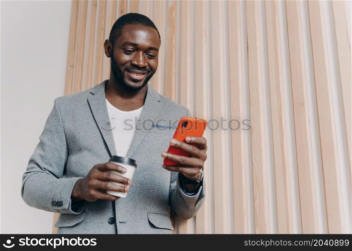 Enjoying coffee break at work. Young happy positive african man office worker in suit using mobile phone and chatting with friends, texting sms or checking email, looking at smartphone and smiling. Enjoying coffee break at work. Young positive african man office worker in suit using mobile phone