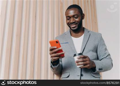 Enjoying coffee break at work. Young happy positive african man office worker in suit using mobile phone and chatting with friends, texting sms or checking email, looking at smartphone and smiling. Enjoying coffee break at work. Young positive african man office worker in suit using mobile phone