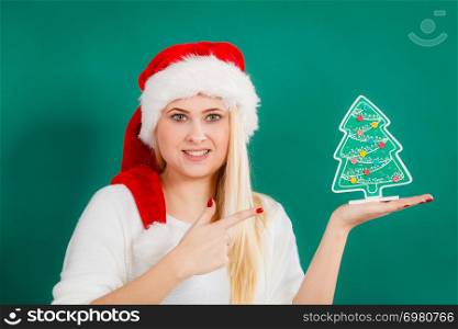 Enjoying cozy holiday interior, seasonal accessories concept. Blonde young woman in Santa hat holding little decorative Christmas tree.. Woman in Santa hat holding little Christmas tree.