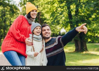 Enjoying good atmosphere. Family harmony. Happy family walk in green park, have happy expressions. Affectionate father points with fore finger into distance as tries to show beautiful sunset