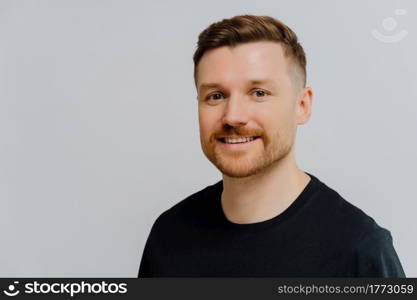 Enjoying life. Close up portrait of happy delighted ginger unshaved man looking at camera with positive face expression while standing in black tshirt against grey wall. People and positive emotions. Handsome unshaven man smiling at camera and expressing positivity
