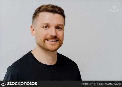 Enjoying life. Close up portrait of happy delighted ginger unshaved man looking at camera with positive face expression while standing in black tshirt against grey wall. People and positive emotions. Handsome unshaven man smiling at camera and expressing positivity