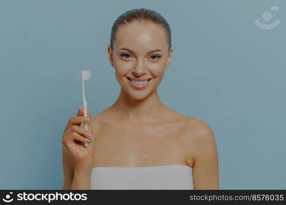 Enjoying morning routine. Happy young woman brushing teeth after shower, smiling female with bare shoulders holding toothbrush, looking at camera, isolated on blue. Oral hygiene and healthy lifestyle. Oral hygiene. Happy young woman brushing teeth after shower, smiling female holding toothbrush