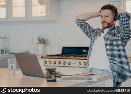 Enjoying remote work. Relaxed bearded male freelancer sitting at table and resting after video meeting with colleagues while using laptop and headset. Freelance and distant work concept. Happy young male worker holding hands behind head and looking at laptop screen at home