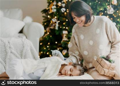 Enjoying xmas at home. Affectionate young mother poses near sleeping daughter, going to make surprise, holds present gifts, believe in miracles. Magic and sweet moment. Family holiday concept
