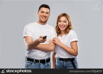 entertainment, television and people concept - portrait of happy couple in white t-shirts with tv remote control over grey background. happy couple with tv remote control