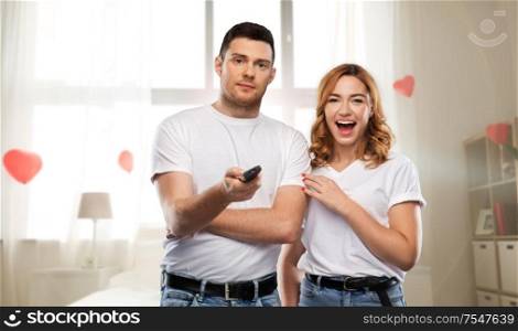 entertainment, television and valentine&rsquo;s day concept - couple in white t-shirts with tv remote control over bedroom decorated with heart shaped balloons background. couple with tv remote control in home bedroom