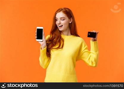 Enthusiastic pretty modern redhead girl advertising mobile banking, shopping online, holding smartphone and credit card, looking curious telephone screen, standing orange background.. Enthusiastic pretty modern redhead girl advertising mobile banking, shopping online, holding smartphone and credit card, looking curious telephone screen, standing orange background