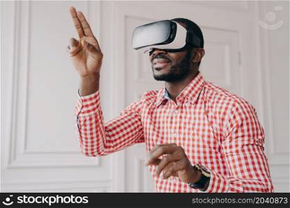 Enthusiastic smiling african ethnicity guy in VR headset glasses enjoying virtual reality online trying cyber world simulation at work, raised hands touching air with fingers, having fun with 3D gadget. Impressed african ethnicity guy in VR headset glasses enjoying virtual reality online at work