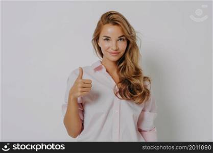 Enthusiastic young Caucasian woman keeps thumb up recommends something very good says yes gestures indoor dressed in pink shirt isolated over grey background shows positive feedback. I like it. Enthusiastic young Caucasian woman keeps thumb up recommends something very good says yes