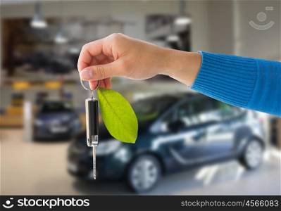 environment, people, transport and ecology concept - close up of hand holding key with green leaf trinket over car show or shop background