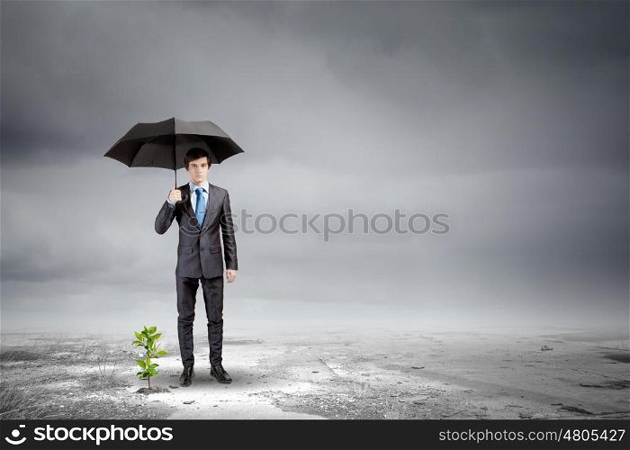 Environment protection. Young businessman protecting little sprout with umbrella