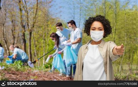 environment, volunteering and charity concept - woman in protective mask pointing finger to camera over group of volunteers working at park on background. volunteer woman in mask pointing finger to camera