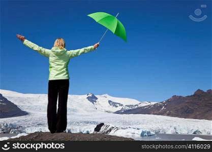Environmental concept shot of a blond woman dressed in green with a green umbrella standing arms outstretched in front of a melting glacier
