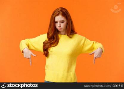 Envy, sad and sulky cute redhead woman in yellow sweater, looking and pointing down with jealousy or disappointment, feeling jealousy and regret missing chance, standing orange background.. Envy, sad and sulky cute redhead woman in yellow sweater, looking and pointing down with jealousy or disappointment, feeling jealousy and regret missing chance, standing orange background