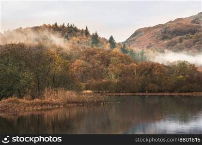 Epic Autumn landscape image of River Brathay in Lake District lookng towards Langdale Pikes with fog across river and vibrant woodlands