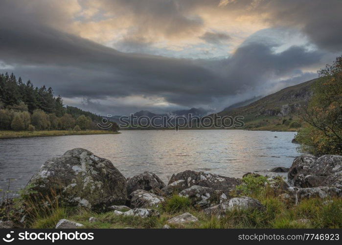 Epic Autumn landscape image of Snowdon Massif viewed from shores of Llynnau Mymbyr at sunset with dramatic dark sky and clouds