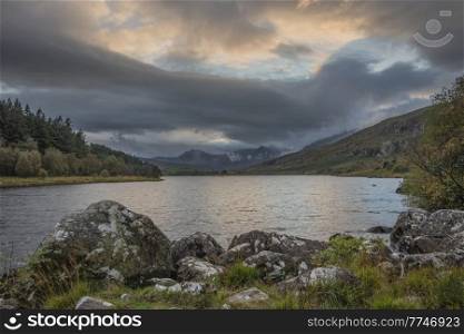 Epic Autumn landscape image of Snowdon Massif viewed from shores of Llynnau Mymbyr at sunset with dramatic dark sky and clouds