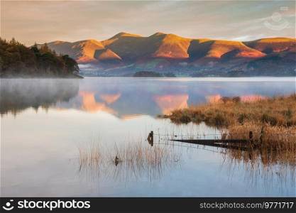 Epic Autumn sunrise landscape image looking from Manesty Park in Lake Distict towards sunlit Skiddaw Range with mit rolling across Derwentwater surface
