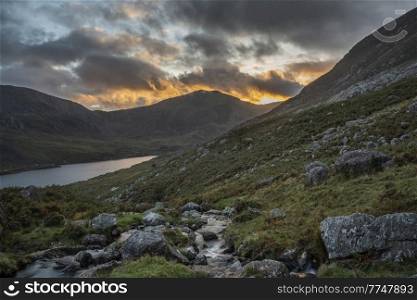 Epic dramatic Autumn sunset landscape image of Llyn Ogwen and Tryfan in Snowdonia National Park with stream and rocks in foreground