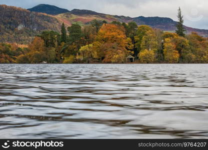 Epic landscape image of boathouse on Derwentwater in Lake District surrounded by vibrant Autumn color woodland