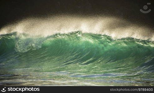 Epic landscape image of jade turquoise waves crashing onto shore and rocks in Kynance Cove Cornwall with glowing sunrise background and water spray droplets in wind