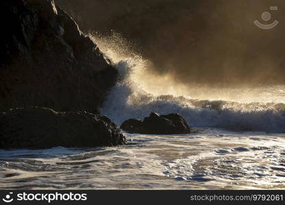 Epic landscape image of jade turquoise waves crashing onto shore and rocks in Kynance Cove Cornwall with glowing sunrise background and water spray droplets in wind