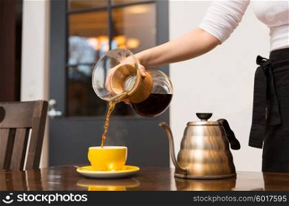 equipment, coffee shop, people and technology concept - close up of barista woman with glass coffeemaker and pot pouring hot coffee to cup at cafe bar or restaurant kitchen