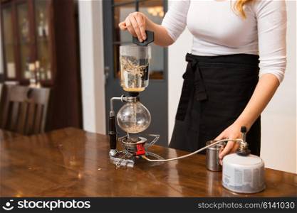 equipment, coffee shop, people and technology concept - close up of woman with butane gas burner heating water in siphon coffeemaker at cafe bar or restaurant kitchen