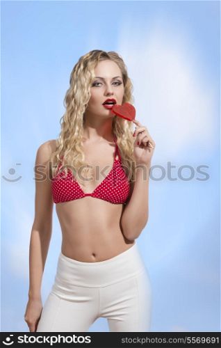erotic blonde woman taking romantic heart shaped lollipop and posing with sexy bikini, long blonde curly hair and white pants.