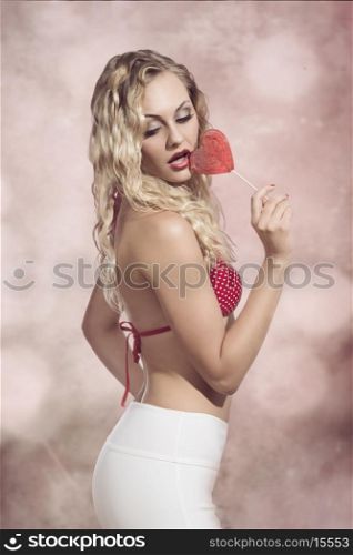 erotic summer blonde girl posing with bikini and sexy pants, eating heart shaped lollipop. Sensual style
