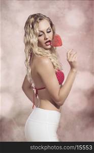 erotic summer blonde girl posing with bikini and sexy pants, eating heart shaped lollipop. Sensual style