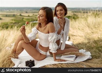 Erotic women in lingerie poses with lollipops on blanket in the field. Female persons with slim body in underwear leisures on meadow, relaxation on sunset, feeling of freedom. Women in lingerie poses with lollipops in field