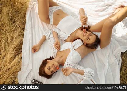 Erotic women in lingerie poses with lollipops on blanket in the field, top view. Female persons with slim body in underwear leisures on meadow, relaxation on sunset, feeling of freedom. Women in lingerie poses with lollipops in field