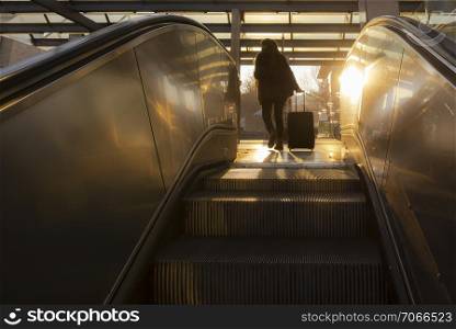 Escalators stairs in a modern city at sunrise and the silhouette of a young woman traveler with a trolley bag