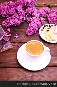 Espresso coffee in a white cup with a saucer, behind a blooming bouquet of lilac