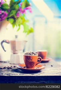 Espresso cup full of coffee beans on kitchen table with coffee pot on background of terrace,sky and flowers