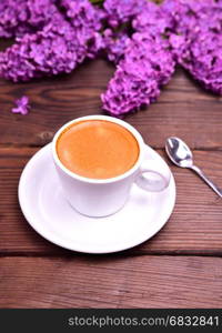 Espresso in a white cup with saucer and spoon, behind a bouquet of purple lilac, top view